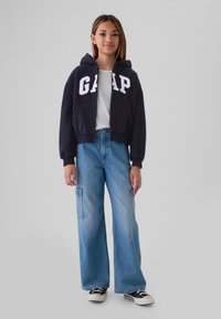 Mörkblå huvtröja med vit "GAP"-logotyp, kombinerad med ljusblå vida jeans med cargofickor och svarta sneakers med vita sulor.
