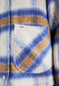 Kockovaná košeľa vyrobená z mäkkej látky v modrej, hnedej a bielej farbe. Obsahuje náprsné vrecko s nálepkou "Lee" a okrúhle biele gombíky.
