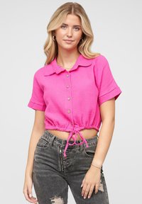 Kortermet, cropped knapper-up skjorte i lys rosa med krage, snøring i livet og teksturert stoff. Kombinert med slitte grå jeans.