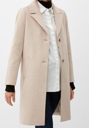 Manteau en laine beige avec col à revers cranté, poches latérales et fermeture à boutons, porté sur une chemise blanche et un pull à col roulé noir.