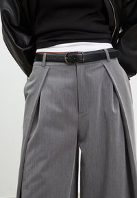 Sive hlače s pliseom izrađene od teksturirane tkanine, opremljene crnim remenom sa srebrnom kopčom. Nosite ih s prilagođenim gornjim dijelom i kožnom jaknom.