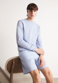 Felpa di cotone azzurro chiaro con colletto a giro e polsini a coste abbinata a pantaloni corti azzurro chiaro. Modello seduto su una sedia curva di legno.