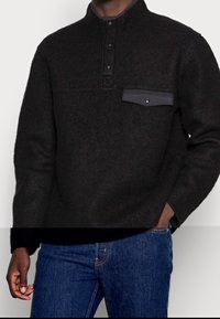 Mustapintainen fleece-poolopaita, jossa on neljännesvetoketju, nappikiinnitys ja rintatasku tummalla yksityiskohdalla. Yhdistetty sinisiin farkkuihin.