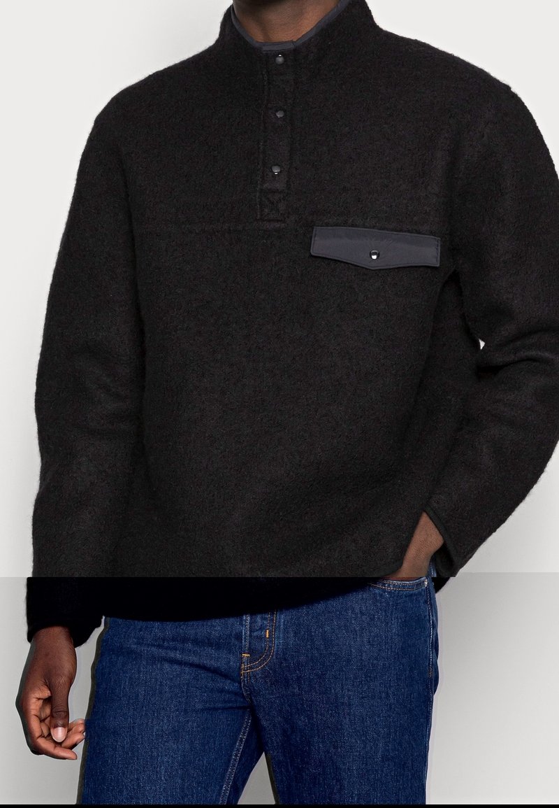 Mustapintainen fleece-poolopaita, jossa on neljännesvetoketju, nappikiinnitys ja rintatasku tummalla yksityiskohdalla. Yhdistetty sinisiin farkkuihin.