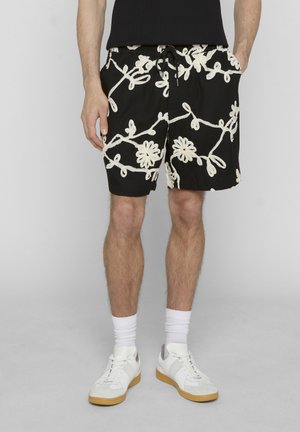Mand iført sorte shorts med hvid blomsterbroderi, hvide sokker, hvide sneakers, stående mod grå baggrund.