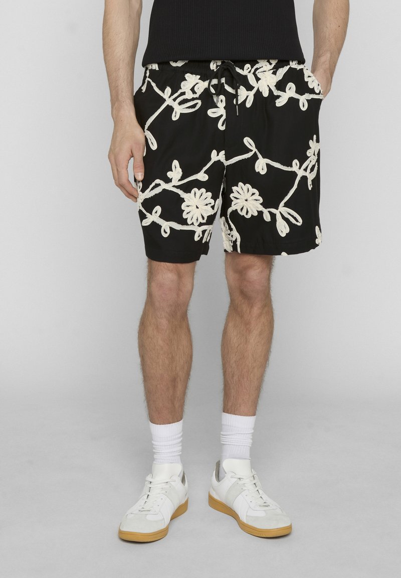 Homme portant un short noir avec une broderie florale blanche, des chaussettes blanches, des baskets blanches, debout devant un fond gris.