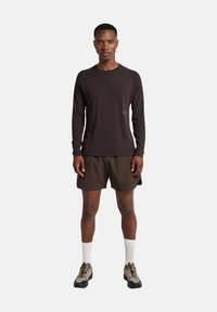 Mann steht in einem dunklen Langarmhemd, dunklen Shorts, weißen Socken und beigen Wanderschuhen vor einem schlichten hellen Hintergrund.
