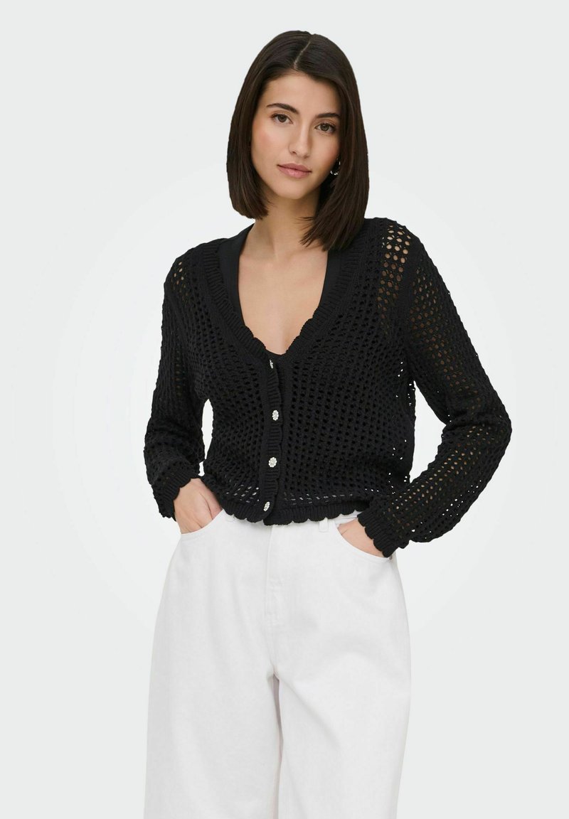 Mujer con cabello liso y oscuro viste un cárdigan negro de punto abierto con botones y pantalones blancos de tiro alto, con las manos en los bolsillos, fondo neutro.