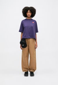 T-shirt viola a maniche corte e cropped con un piccolo logo rosso; abbinata a pantaloni a palazzo beige e un cappellino nero. Sneakers nere completano il look.