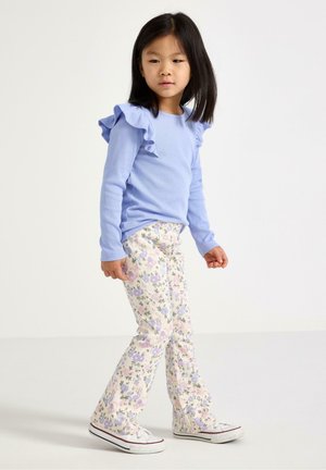 Ung pige med skulderlangt hår iført en lyseblå bluse med flæser, blomstrede bukser og hvide sneakers, stående mod en ensfarvet baggrund.