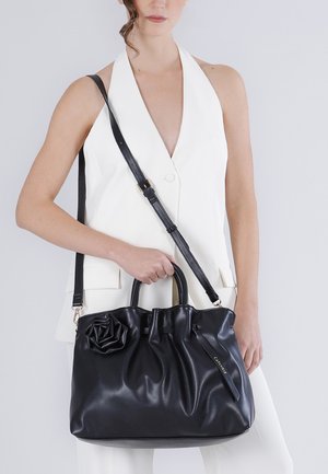 Donna con blazer bianco senza maniche che tiene in mano una grande borsa a tracolla nera in pelle con dettaglio floreale e tracolla regolabile.