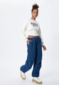 Witte cropped sweatshirt met meerkleurige graphics; marineblauwe losse broek; witte platformsneakers met tan kleurige zolen; creolen.