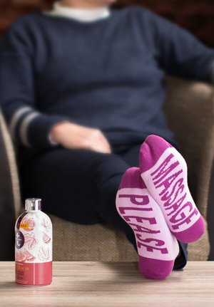 Purple and pink socks with "MASSAGE PLEASE" text, made of soft fabric. A bottle of body wash is placed on a wooden table nearby.