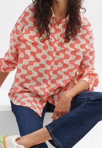 Shirt features a pink and cream wave pattern, long sleeves, button-down front, and two chest pockets. Paired with dark blue jeans.