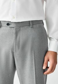 Pantalones grises a medida hechos de tela texturizada, con frente plano, dos bolsillos laterales y cierre de botón en la cintura.