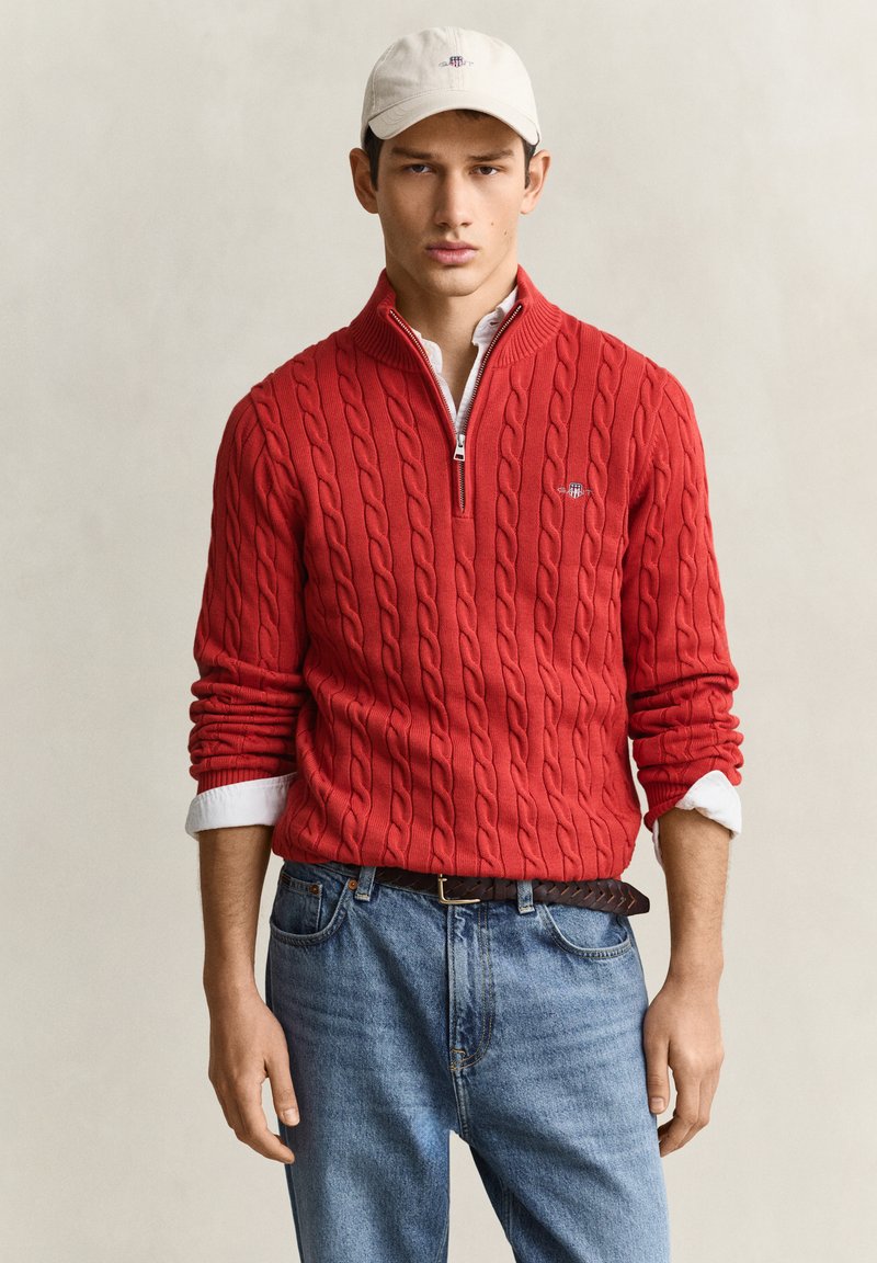 Jeune homme portant un pull rouge à torsades avec une demi-fermeture éclair, un jean bleu, des poignets de chemise blancs et une casquette de baseball blanche sur un fond uni.