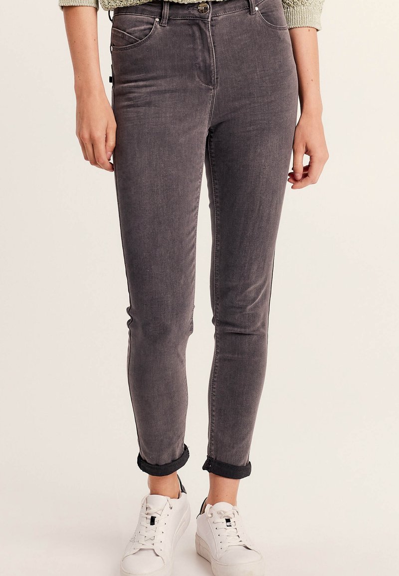 Jeans skinny in denim grigio con vita media, cinque tasche e orli arrotolati, abbinati a sneakers bianche. La texture appare liscia.