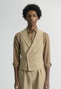 Chaleco beige de lino de doble botonadura con un diseño estructurado, que presenta dos bolsillos delanteros y una textura suave. Estilizado con una camisa marrón.