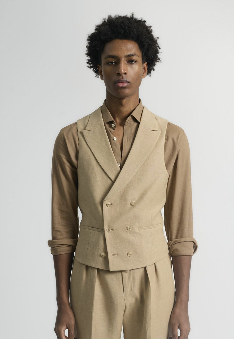 Chaleco beige de lino de doble botonadura con un diseño estructurado, que presenta dos bolsillos delanteros y una textura suave. Estilizado con una camisa marrón.