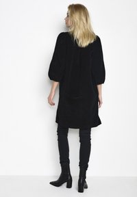 Abito in velluto a coste nero con una vestibilità ampia e maniche a sbuffo, abbinato a jeans neri aderenti e stivali alla caviglia. Texture liscia e design semplice.