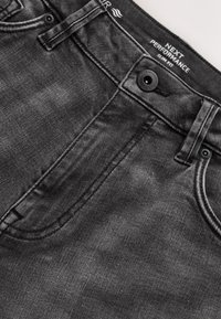 Jean slim gris foncé montrant un bouton, des passants de ceinture, une poche avant et une étiquette "Next Performance Slim Fit" à l’intérieur de la ceinture.