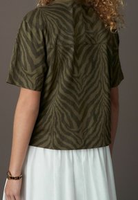 Kobieta w oliwkowej, krótkiej bluzce w ciemny wzór zebry i białej spódnicy, stojąca na tle jednolitego tła.