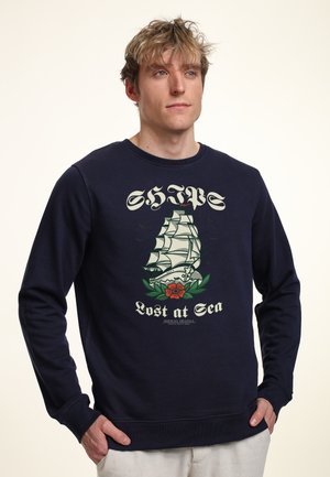 Felpa navy con grafica di una nave a vela accompagnata da un fiore e testo sottostante. Realizzata in tessuto morbido con polsini e orlo ribattuti.