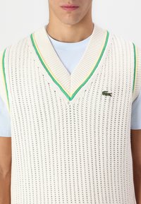 Pleteni bezrukavni prsluk u boji off-white s V-izrezom, koji ima zelene i žute pruge oko ovratnika i izvezeni logotip.