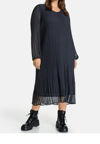 Černé plisované midi šaty s dlouhými průhlednými rukávy a kulatým výstřihem, doplněné o robustní černé šněrovací boty. Hladká textura, elegantní design.