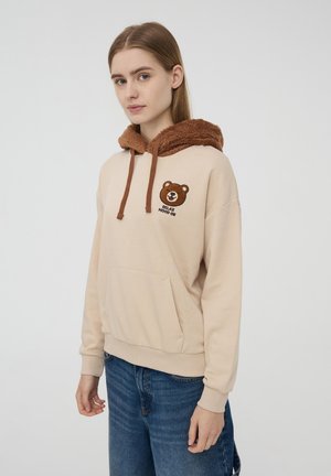 Felpa beige con un cappuccio marrone e peloso. Presenta una grafica di un orso e la scritta "RELAX" sul petto. Tasca anteriore a marsupio, polsini e orlo a coste.