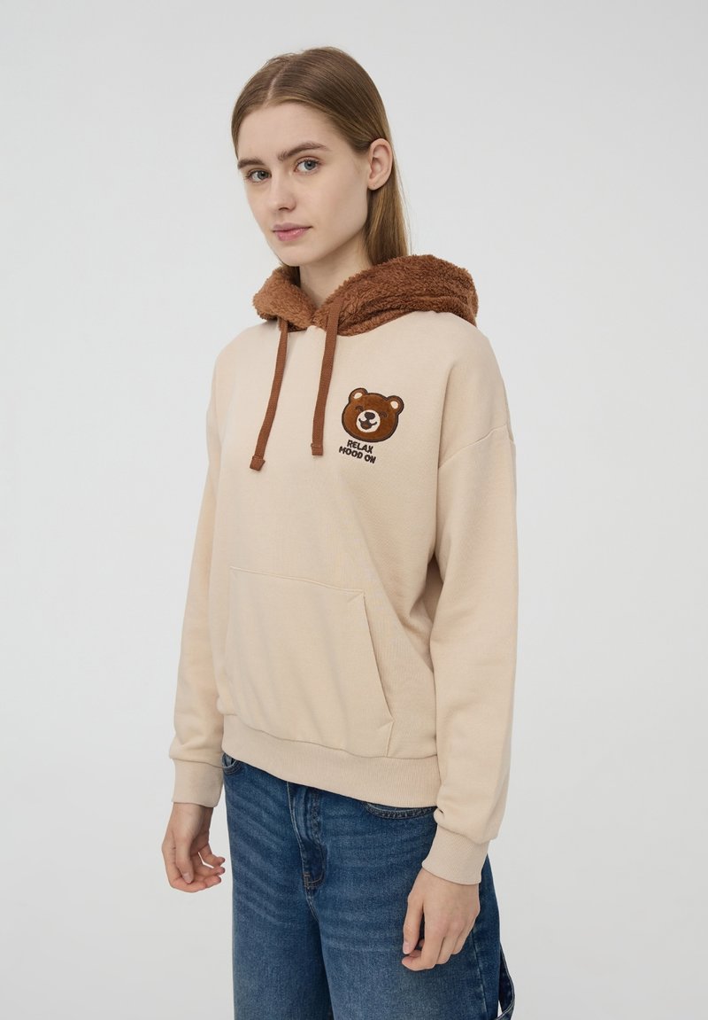 Felpa beige con un cappuccio marrone e peloso. Presenta una grafica di un orso e la scritta "RELAX" sul petto. Tasca anteriore a marsupio, polsini e orlo a coste.