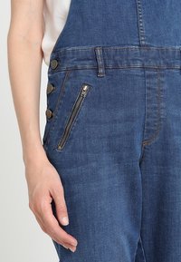 Denimbyxor med hängslen i en mörkblå färg, sidoknappar och en framfick med dragkedja i brons. Slät, strukturerad tyg.