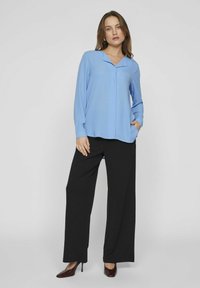 Lyseblå langermet bluse med V-hals, kombinert med svarte vide bukser og mørkebrune hæler. Glatt stoff og avslappet passform.