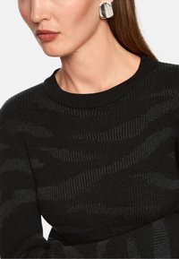 Black knitted sweater with textured stripes and a ribbed neckline, paired with a metallic silver square earring.