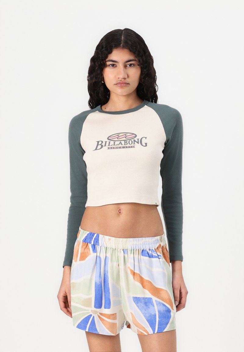 Crop top con maniche raglan in beige e verde con logo stampato "Billabong". Abbinato a shorts colorati con fantasia, in blu, arancione e verde.
