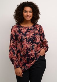 Blouse florale en marine foncé avec des motifs de points roses et oranges. Présente des manches longues avec des poignets smockés et un col rond. Texture lisse.