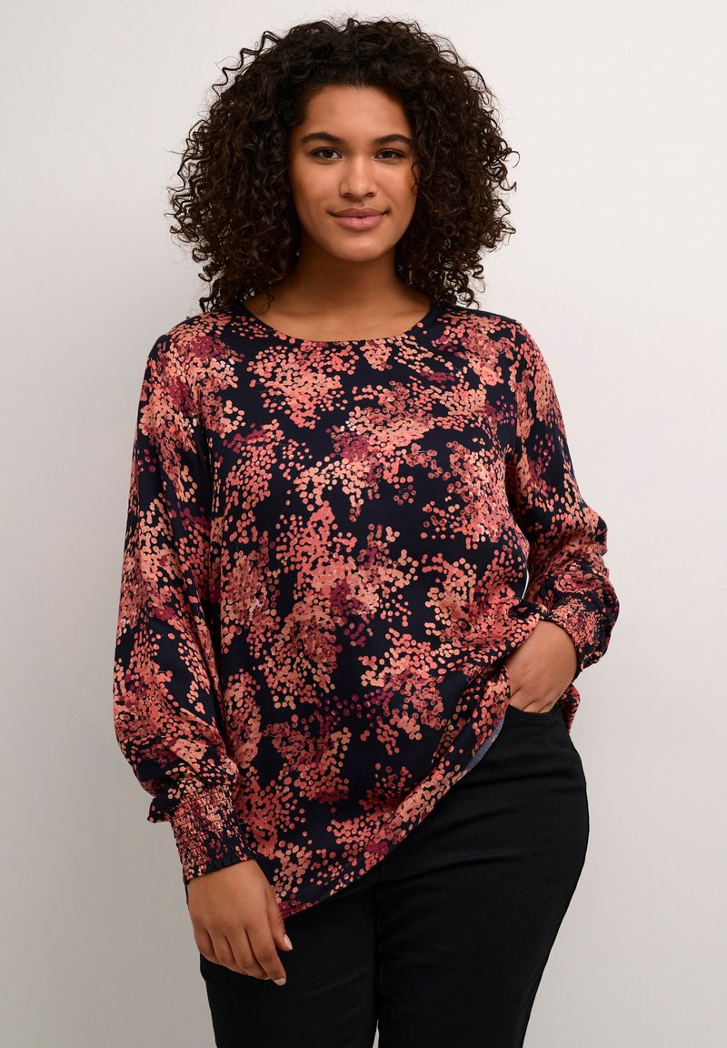 Blouse florale en marine foncé avec des motifs de points roses et oranges. Présente des manches longues avec des poignets smockés et un col rond. Texture lisse.