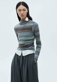 Randig turtleneck-tröja i flera färger, blå, brun och grå, bärs över en vit skjorta med krage och matchas med lösa mörkgrå byxor.