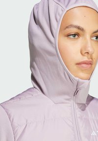 Giacca a vento viola chiaro con cappuccio e zip, caratterizzata da una texture di tessuto morbido e un design imbottito. Il cappuccio aderente incornicia il viso.