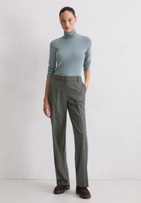 Frau in einem hellblauen Rollkragenpullover, der in grau maßgeschneiderte Hosen gesteckt ist, und braunen Schuhen, steht mit einer Hand in der Tasche gegen eine weiße Wand.