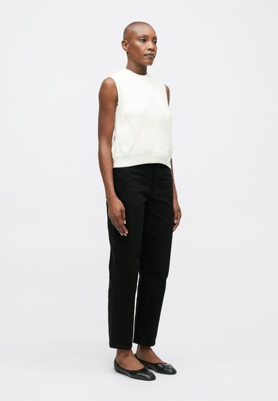 Mujer con cabeza rapada, vistiendo una camiseta de punto sin mangas blanca, pantalones rectos negros y zapatos planos negros, de pie contra un fondo blanco.