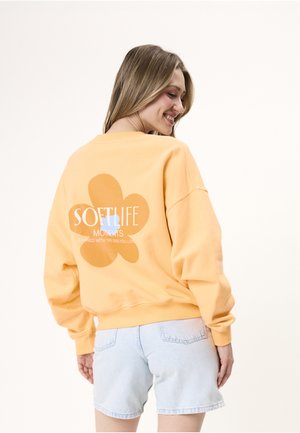 Ung kvinde smiler, iført gul sweatshirt med stort blomsterbillede og teksten "Soft Life Moments" samt lyse denimshorts, vender ryggen til.