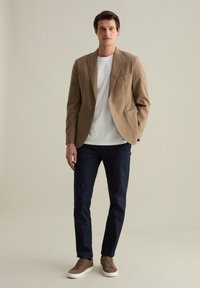 Blazer beige sopra una maglietta bianca, abbinato a pantaloni blu scuro e sneaker marroni. Il blazer ha due tasche frontali e una vestibilità su misura.