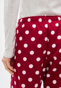 Άτομο που φορά κόκκινο παντελόνι με λευκές πουά και ανοιχτογκρι μακρυμάνικο τοπ, με το χέρι χαλαρό δίπλα στο σώμα.