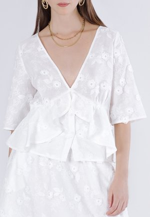 Nainen pukeutuneena valkoiseen kukkakirjailtuun paitaan, jossa on röyhelöt, puolipitkät hihat, syvä V-kaula-aukko ja kultaiset rengaskorvakorut, joissa on kerrokselliset ketjut.