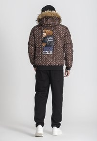 Giacca puffer marrone con cappuccio in pelliccia sintetica, che presenta una grafica di un orso sul retro. Il design include un motivo a monogramma e polsini a coste.