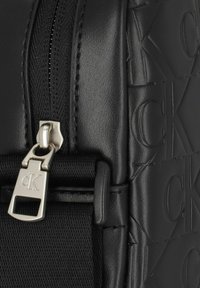 Borsa nera con texture e motivo a rilievo "CK", dotata di una cerniera argento e di un robusto dettaglio in tessuto. Finitura liscia e design elegante.