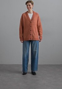 Knitted rust-orange cardigan with a V-neck, front buttons, and ribbed cuffs, paired with loose blue denim jeans and black shoes.