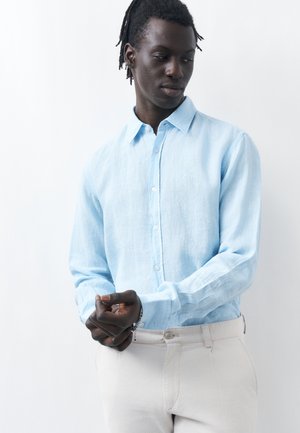 Férfi világoskék gombos ing mandzsettáját igazítja, világos színű nadrágot visel, sima fehér háttér előtt.