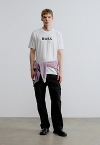 Valge T-särk, millel on "BOSS" logo, lavendlivärvi kampsun, mis on vöökohal seotud, mustad cargo-püksid ja mustad jalanõud, esitletud modellil ühevärvilise tausta ees.